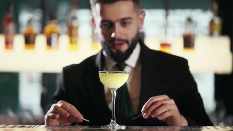 Professional barman in black suit and tie neatly decorating tasty alcoholic cocktail using small clothespin and twig of lavender. Concept of occupation, restaurant servings and unique recipe.