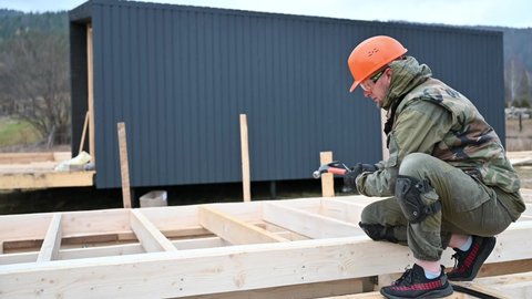 Man worker building wooden frame house on pile foundation. Carpenter hammering nail into wooden board, using hammer. Carpentry concept.