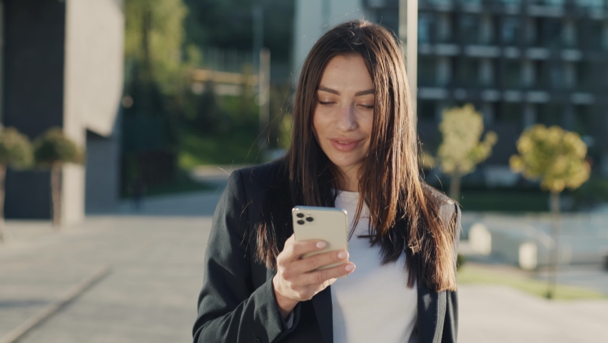 Portrait young attractive woman walking using the phone in urban city at sunlight look around feel happy. Portrait girl with mobile. Slow motion