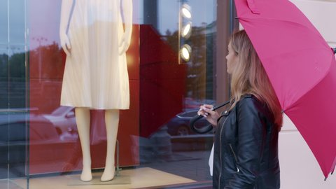Blonde stands near fashion store, looks at shop-window with mannequins in fashionable clothes. Beautiful girl wants to update her wardrobe, go shopping in city. Young woman admires dress at street
