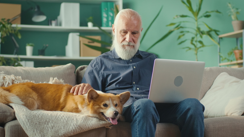 Busy Senior Man Uses Laptop Computer, While Sitting on a Couch with His Lovely Pet in the Light Living Room. Happy Mature Man is Working from Home with Pet Friend.Home Office and Happy Life Concept. - Powered by Shutterstock - Get 15% off with code: PIKWIZARD15