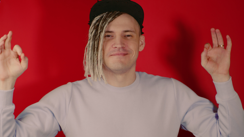 Young handsome smiling man in black cap meditating, looking at camera on red background in studio. Blonde guy in violet sweatshirt keeping hands in zen gesture, trying to relax.