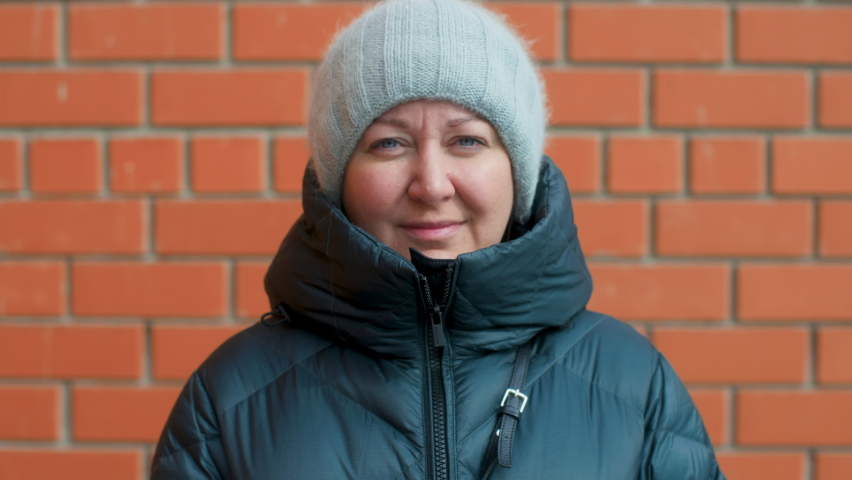 Portrait of caucasian chubby woman in dark green jacket and winter hat standing against red brick wall background, looking at camera and around. Close-up