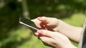 Teenager Girl Uses Cell Phone In Summer In The Park, Close-up Of Hands And Smartphone, Side View. Teenager Communicates with Friends in Social Networks, Watches Video Content, Writes SMS.