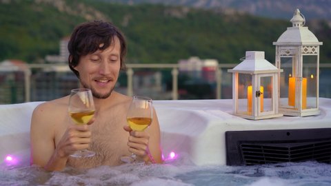 A funny young man is having a good time in the hot tub on a rooftop. He drinks wine from two glasses. She did not come and he is happy