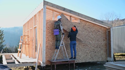 Carpenters mounting wooden OSB board on the wall of future cottage. Men workers building wooden frame house on pile foundation. Carpentry and construction concept.