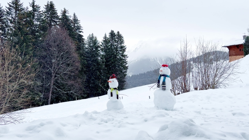 Two snowman built dressed with hat, scarf over mountain panorama and forest at winter during snowfall