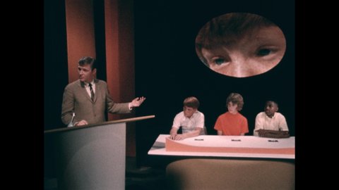 1960s: Children sit at table with an image of a girl's face above them. Man stands at podium and talks. Boy sits at table, talks. Children roll their eyes, put head in hands, press a button.