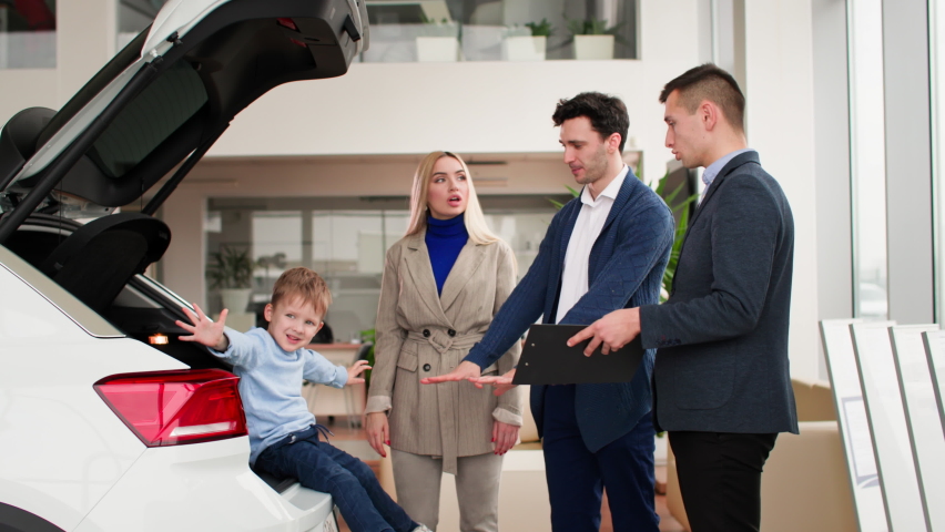 Buying a automobile, happy young parents with son in trunk choose new car for their family together with sales manager in auto center