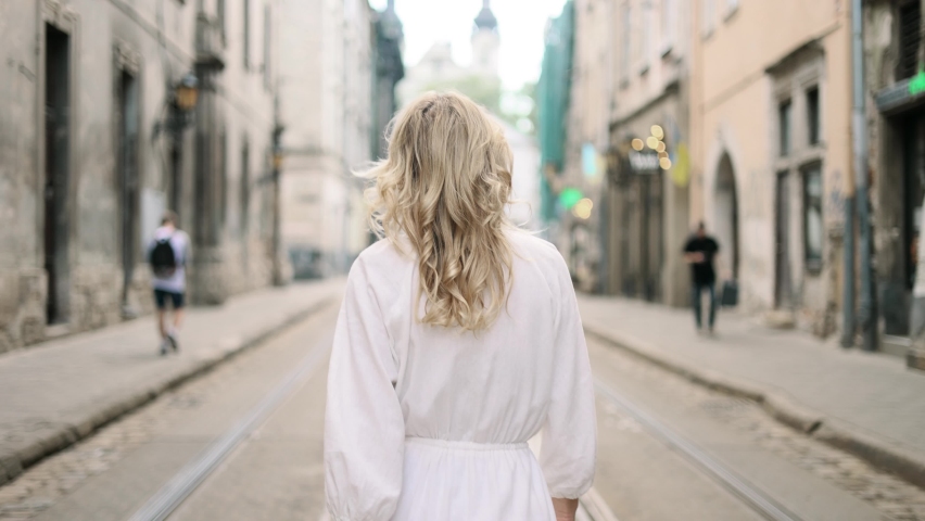 Attractive middle aged woman in white dress walking down the street turns around with flying hair and looking at ahead outdoors Happy relaxed lady walking on the city centre enjoying beautiful day Royalty-Free Stock Footage #1090896165