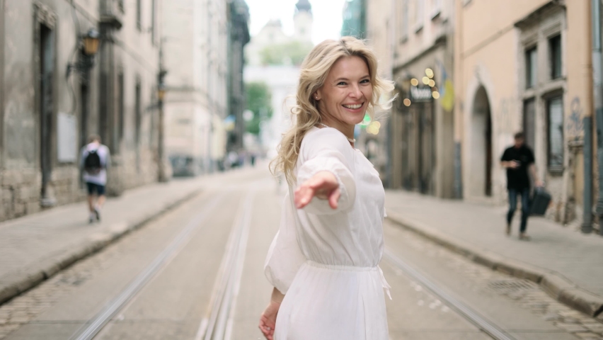 Attractive middle aged woman in white dress walking down the street, turns around with flying hair and looking at the camera. Happy relaxed lady walking on the city centre enjoying. | Shutterstock HD Video #1090896165