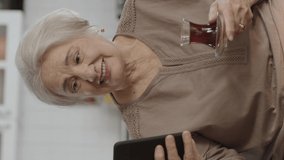 Happy old grandmother holding mobile phone, video call with loved ones while drinking tea. An elderly woman learning to use modern technology device. Video for the vertical story.