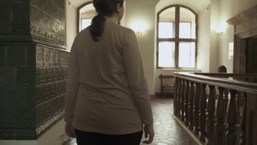 Overweight young woman looks around antique room with tiled stove and wooden railings. Lady comes to big old window, looks out of it, rear view. Wooden benches stand near wall. Medieval room interior.