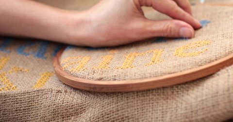 POV hands hold needle and embroider cross stitch letter n with blue thread on burlap in embroidery hoop. Yellow embroidered text - Ukraine. Traditional needlework in blue yellow colors on canvas