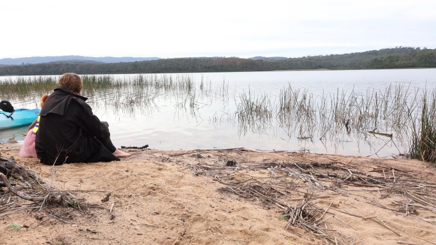 A man in a big rain jacket sits on the shore of a lake with his daughter in a ballet dress.