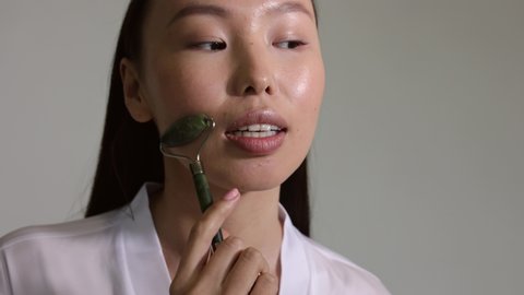 Young asian girl doing face massage with stone roller. Closeup of ideal beautiful woman face with big lips and beautiful smile. Hands doing face massage with roller. Self care and self treatment