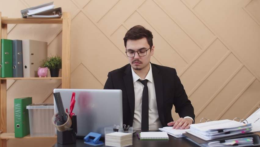 Office worker gets very upset by lifting several pages of working documents, front view. Man in an office suit grimace lifting sheets from a stack of working documents