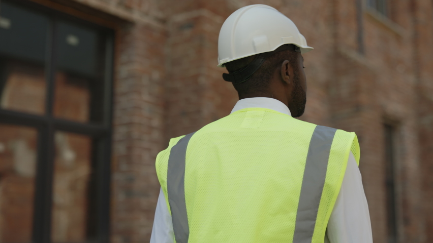 Back view of african american male builder in hardhat walking along wall of unfinished building. Competent architect examining and controlling working process at construction site.