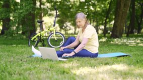 senior retired sports woman workout training fitness exercises or yoga outside using laptop online lessons outdoors in park. Active mature female on the mat practicing yoga, stretching on the lawn
