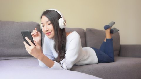 A young attractive woman lies on the couch as she listens to music from her smartphone through her Bluetooth headphones. 