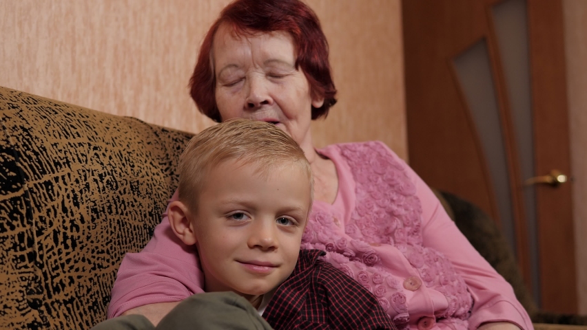 Close-up of a happy grandmother kissing her eight-year-old grandson at home sitting on the couch. The grandson came to visit an elderly grandmother.