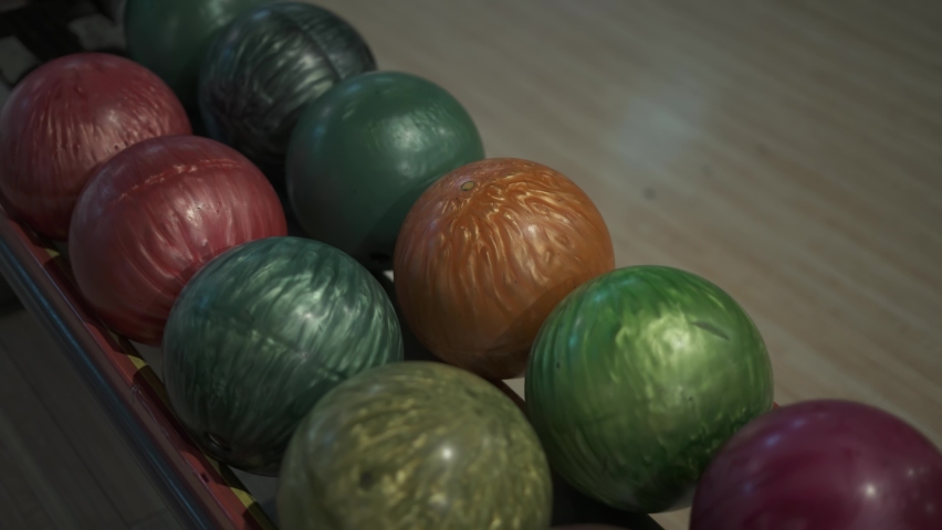 Unrecognizable woman approaches the rollout system in bowling alley and takes heavy orange ball with both hands, close-up. An amateur plays a sports game. Variety of multi-colored balls lie in row. Royalty-Free Stock Footage #1090918809