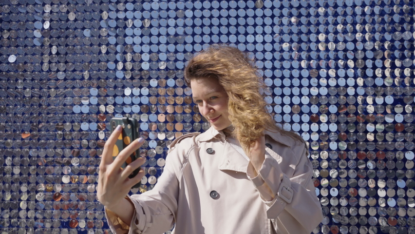 Beautiful woman in beige coat makes selfie at shiny mirror pieces wall during stroll. Young chestnut woman makes photos on sunny day taking city walk