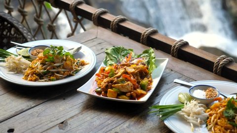 Traditional thai food in outdoor cafe with nature waterfall background. Fried noodle with prawn pad thai and chicken cashew nut on wooden table in restaurant. Asian style food lunch