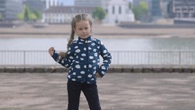 Lovely girl dancing on a lawn with beautiful river in the background. Kid playing with her hair while enjoying warm morning on a shore of city pond. High quality 4k footage