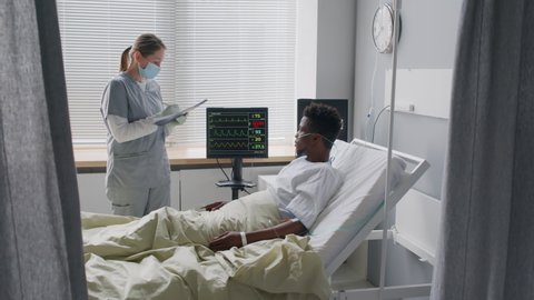 Female medical worker in scrubs and face mask explaining vital signs on patients bedside monitor during checkup in hospital