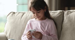 Close up cute little girl play games, watch videos using cellphone seated on sofa alone at home. Young gen Z use modern wireless tech, parental control software app for kids safety on internet concept