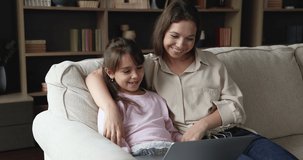 Mother and little 6s daughter watch amusing cartoons on laptop sit on sofa at home. Spend leisure enjoy interesting educational on-line program for kids development, modern technology for fun concept
