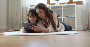 Mom and little daughter spend free time use digital tablet. Cute girl her mum lying on floor on carpet in playroom have fun on internet, hold modern device watch favourite vlog, play videogame concept