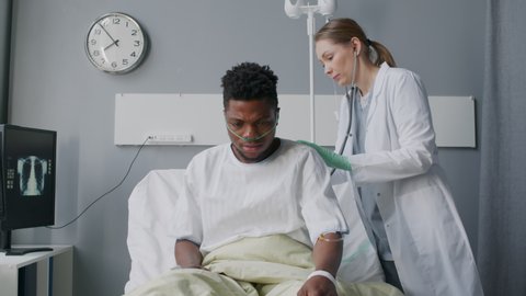 Female doctor using stethoscope on back of young Black man with oxygen tube, lying on bed in hospital bed with his chest xray on bedside monitor
