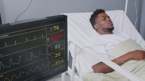 Sick young African American man with IV drip and oxygen tube in his nose lying in hospital bed, looking away. Patient bedside monitor with vital indicators on foreground