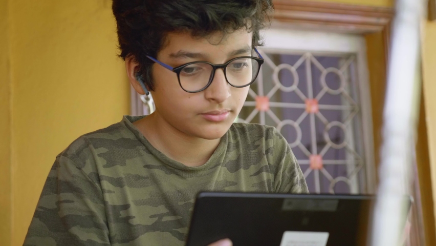 A young Indian Asian male student wearing wireless headphones and eyeglasses reading or interacting during online group class  video activity using a Tablet. Remote school, distance education concept  Royalty-Free Stock Footage #1090943201