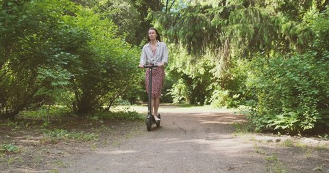 Woman in dress is riding towards the camera along a path in a park with green trees. Eco travel concept, green energy, vehicle. Front view, wide shot, daytime, spring summer.