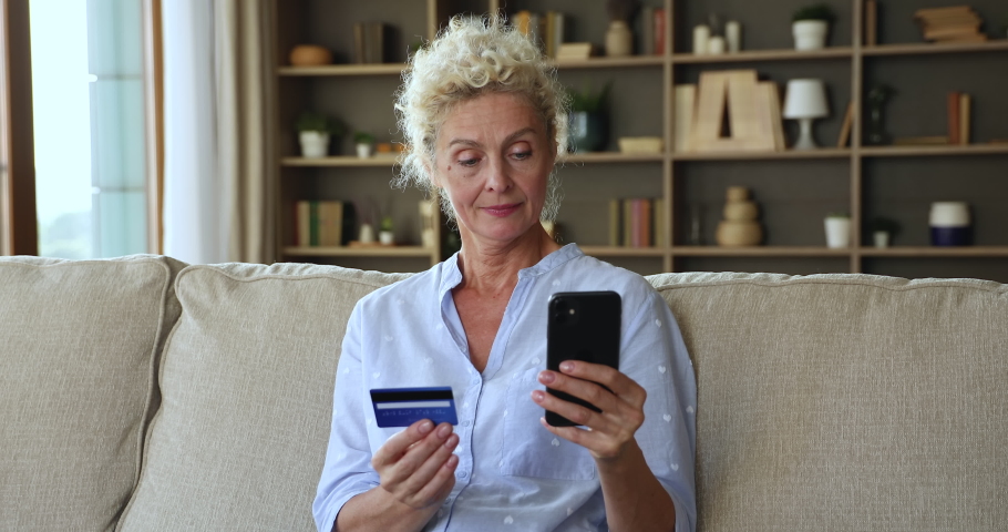 Older 50s woman using cellphone holding credit card try to make payment having problems, due to insufficient funds, password forgot, blocked account. Insecure e-payment, online theft and fraud concept Royalty-Free Stock Footage #1090949451