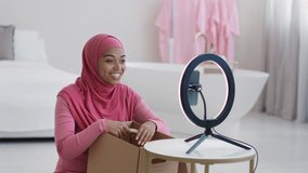 New device unpacking. Young happy muslim black lady wearing headscarf demonstrating wireless headphones to smartphone camera, streaming video online at home, slow motion