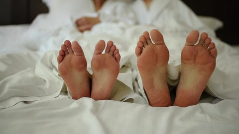 Man scratches his leg with other leg. Bare feet covered with blanket close up. Young couple in love lying in bed in white bedding. Male and female legs bare. Morning newlyweds wedding barefoot