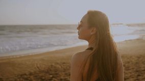 A young woman in sunglasses walks on the beach by the sea at sunset. A strong wind blows her long hair. Barefoot on the sand