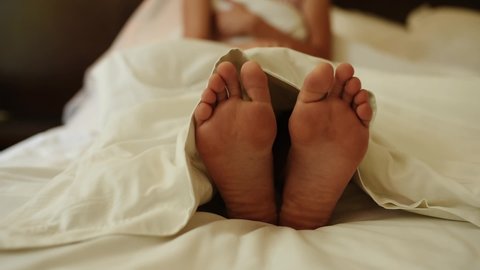 Young girl in bed covered under blanket with bare feet close up. Woman beautiful legs toes macro.  Caucasian woman lifts blanket and bare feet under it. girl covers her bare feet with white blanket