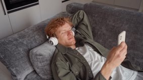 Side view young ginger man in headphone talking on video call using mobile app. Millennial british male freelancer or student lying on sofa with smartphone at home office. Digital communication online
