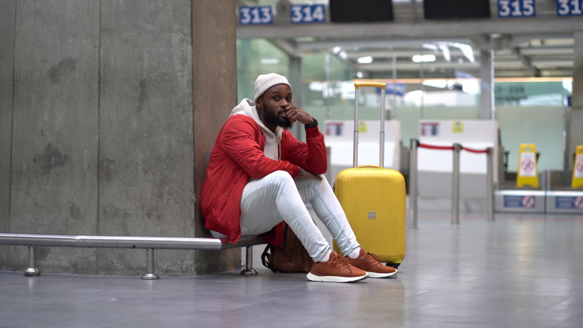 Tired African American man upset at airport his flight is delayed. Nervous traveler male waiting for a plane sitting in empty terminal with baggage. Exhausted guy on a long night connection at airport