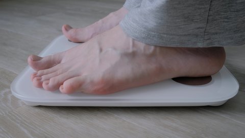 Side view: man weighing himself at home - male bare feet stepping on white digital floor scales: close up. Measuring weight, control, wellness and diet concept
