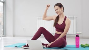 a young woman in a burgundy tracksuit with laptop on a mat in a gym. Record video and online lessons for sports. clothes and shoes for gymnastics. professional trainer. fitness