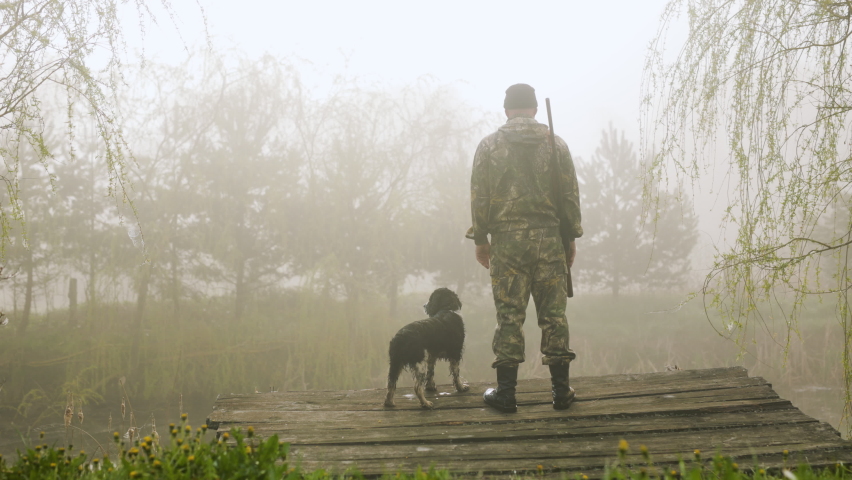 A hunter stands with a dog on a lakeside bridge in fog