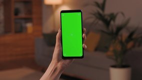 Close up view of female hands holding smartphone with green mock-up screen vertical mode. Woman indoors of cozy home, watching content videos blogs, news or films. Cinematic lighting.