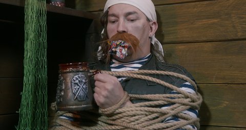 Bound young man with a long red mustache with a gag in his mouth and a bandana on his head. Desperate pirate is holding mug in hand and demanding a drink even while in captivity