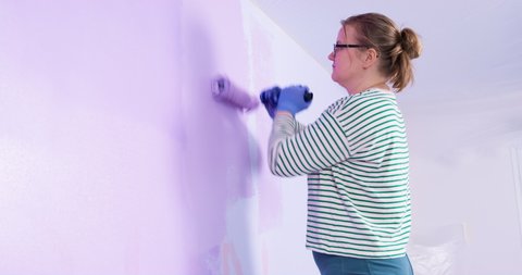 Lady painter carefully paints wall in very peri color , looks closely and removes the speck. Pregnant woman with roller in her hands makes repairs in apartment or children's room